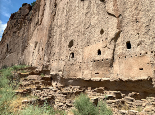 Ancient cliff dwellings with stone structures and small openings, surrounded by greenery and rocky terrain.