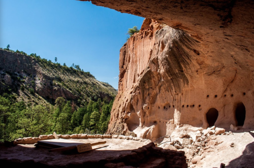 View from a cave dwelling, showcasing rocky cliffs and lush greenery under a clear blue sky.