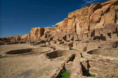 Ancient stone ruins nestled against a rocky cliff under a clear blue sky, with circular structures and grassy areas.