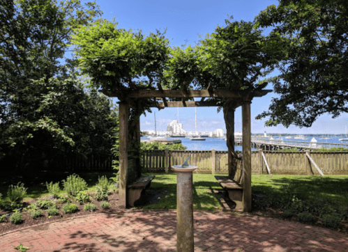 A wooden pergola with greenery overlooks a waterfront, featuring benches and a stone pedestal in a sunny park setting.