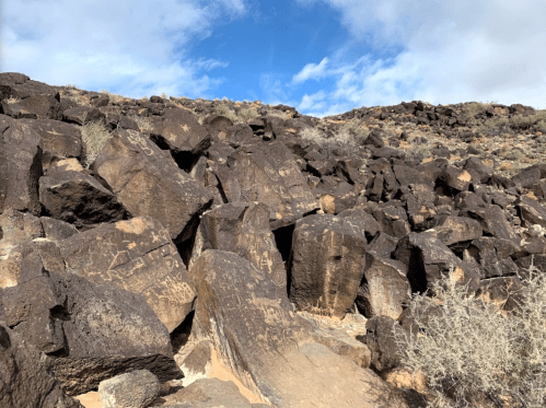 Rocky hillside with dark boulders and sparse vegetation under a partly cloudy blue sky.