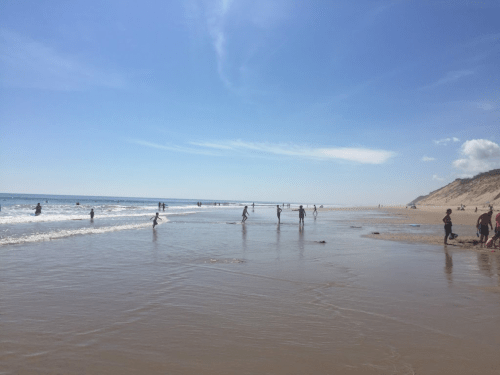 A sunny beach scene with people wading in the water and walking along the shore under a clear blue sky.