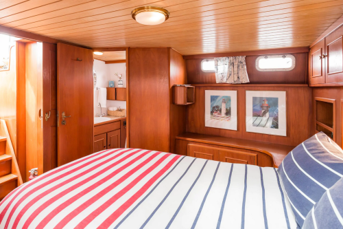 Cozy cabin interior with wooden walls, a striped bedspread, and nautical-themed artwork. Light streams through windows.