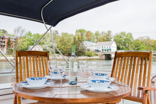A dining table set on a boat with plates, glasses, and a bottle of wine, surrounded by trees and buildings in the background.