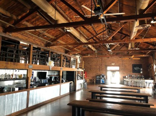 Interior of a rustic bar with wooden beams, metal accents, and long tables, featuring a bar counter and shelves.