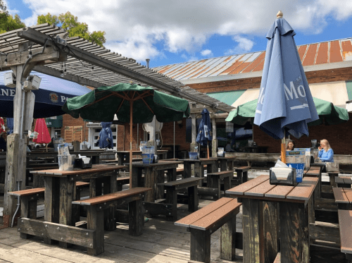 Outdoor dining area with wooden tables, umbrellas, and a rustic atmosphere under a partly cloudy sky.