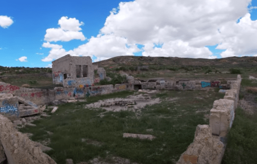 Abandoned stone structure with colorful graffiti, surrounded by grass and hills under a blue sky with clouds.