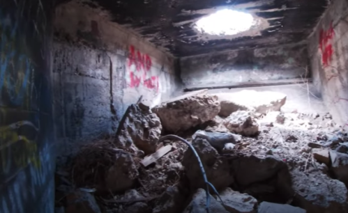 Interior of a dilapidated room with rubble, graffiti on the walls, and a shaft of light coming from a hole in the ceiling.