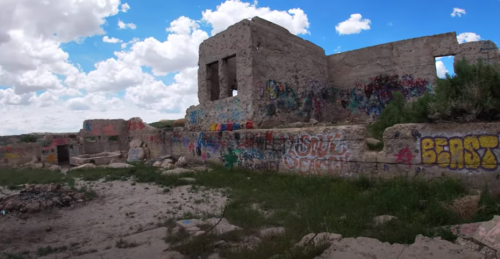 Abandoned building ruins covered in colorful graffiti, surrounded by grass and under a blue sky with clouds.