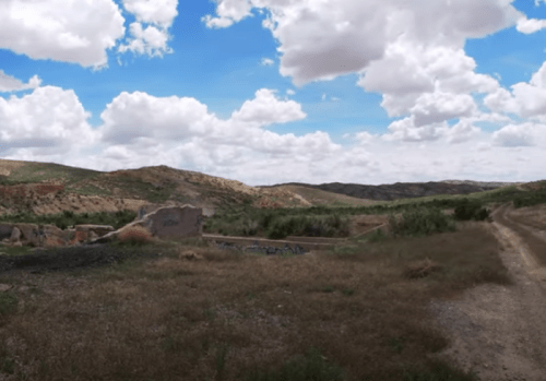 A scenic landscape featuring rolling hills, scattered clouds, and remnants of a structure in a grassy area.
