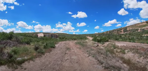A dirt path leads through green shrubs under a bright blue sky with fluffy white clouds in a rural landscape.