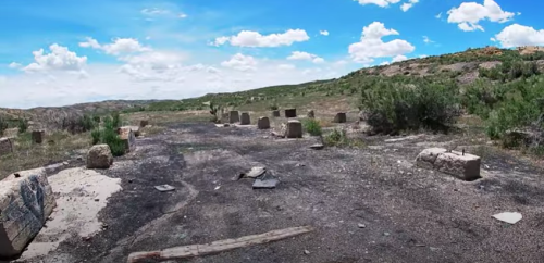 A barren landscape with scattered stone remnants and sparse vegetation under a bright blue sky with clouds.