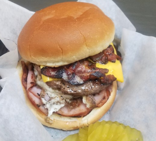 A hearty burger with bacon, cheese, and ham, served on a soft bun with pickles on the side.