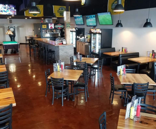 Interior of a casual dining bar with wooden tables, black chairs, a bar area, and multiple TVs showing sports.