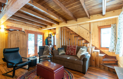 Cozy living room with wooden beams, a sofa, armchair, and a staircase, featuring warm lighting and rustic decor.