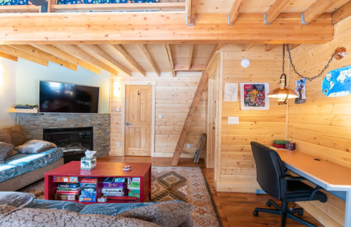 Cozy wooden living room with a TV, stone fireplace, games on a table, and a desk in a bright, inviting space.