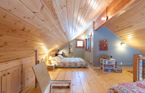 Cozy attic bedroom with wooden walls, two beds, and colorful decor, featuring natural light and storage options.