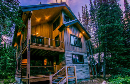 A modern wooden cabin surrounded by tall trees, featuring multiple balconies and warm lighting at dusk.