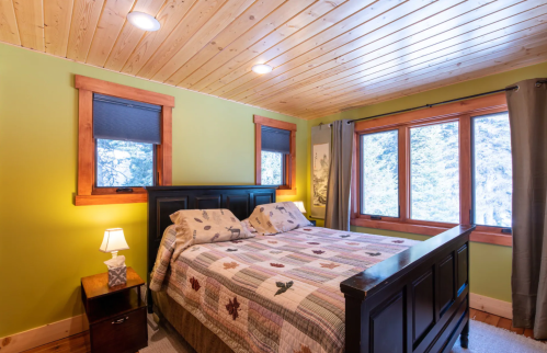 Cozy bedroom with a wooden ceiling, green walls, a quilted bedspread, and large windows overlooking a snowy landscape.