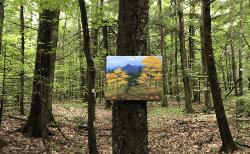 A painting of autumn trees hangs on a tree in a lush green forest, surrounded by tall trees and fallen leaves.