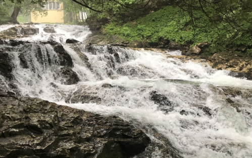 A cascading waterfall flows over rocky terrain, surrounded by lush greenery and a hint of a building in the background.