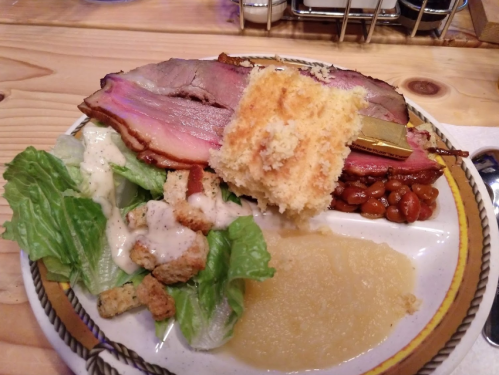 A plate with sliced smoked meat, cornbread, baked beans, lettuce salad with croutons, and applesauce.