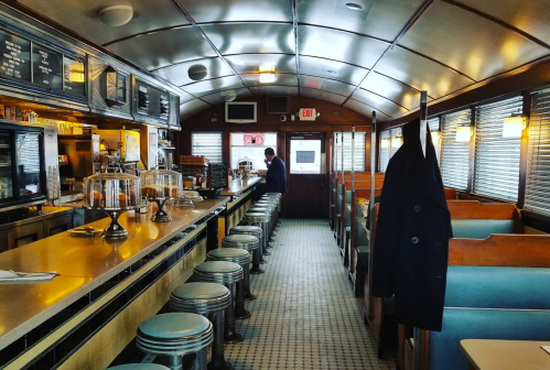 A vintage diner interior with a long counter, stools, and booths, featuring a man standing at the counter.
