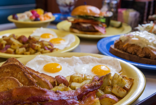 A colorful breakfast spread featuring eggs, bacon, potatoes, and a burger on a table with fruit in the background.