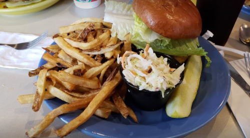 A plate with a burger topped with lettuce, a side of fries, coleslaw in a cup, and a pickle slice.