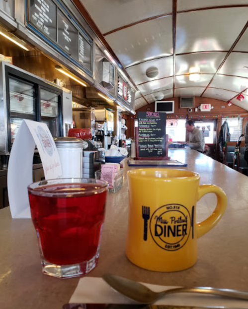 A diner counter with a yellow coffee mug and a glass of red drink, featuring a menu board in the background.