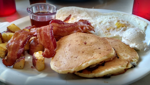 A plate of breakfast featuring pancakes, bacon, eggs, and seasoned potatoes with syrup on the side.