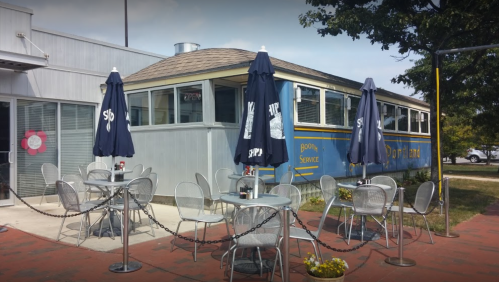 A retro diner with outdoor seating, featuring blue and yellow decor and umbrellas, set in a sunny area.