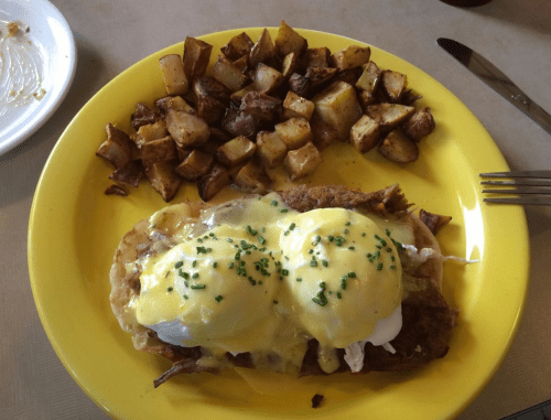 A yellow plate with two eggs Benedict on a tortilla, topped with hollandaise sauce, and a side of roasted potatoes.