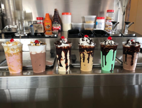 A row of six colorful milkshakes topped with whipped cream and cherries, displayed on a counter with various toppings.
