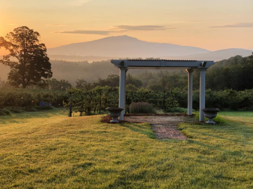 A serene landscape at sunrise featuring a pergola, lush greenery, and distant mountains under a soft, colorful sky.