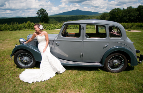 A bride in a white gown sits on a vintage gray car, with a scenic mountain backdrop and lush greenery.