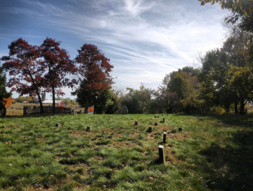 A serene graveyard with grassy grounds, scattered headstones, and colorful autumn trees under a blue sky.