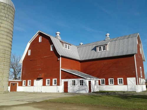 A red barn with a gray roof and white trim, next to a silo, set against a clear blue sky.