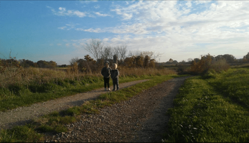Two children walk along a dirt path in a grassy field under a blue sky with scattered clouds.