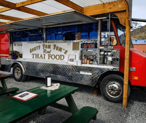 A colorful food truck named "Aunty Yum Yum's Real Thai Food" parked under a wooden shelter with picnic tables.