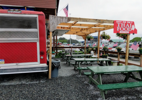 A red food truck with a sign reading "Thai Food" next to outdoor seating and American flags in a gravel area.