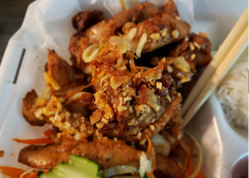 A close-up of crispy chicken wings topped with garlic and fried onions, served with vegetables and chopsticks.