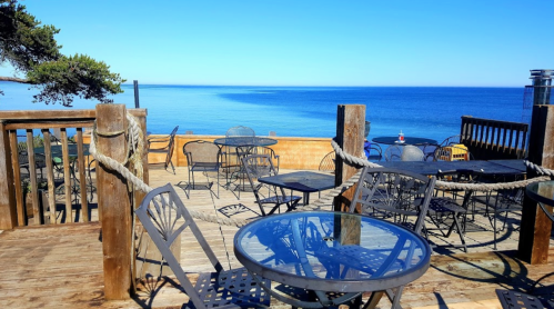 Outdoor seating area with black metal tables and chairs overlooking a calm blue ocean under a clear blue sky.