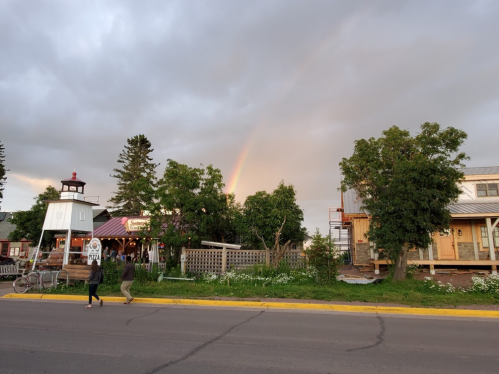 A rainbow arcs over a quaint street scene with trees, buildings, and people walking along the sidewalk.