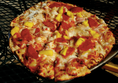 A pizza topped with pepperoni, ham, pineapple, and melted cheese on a round crust, placed on a green wire table.