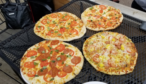 Four pizzas on a black wire rack: one with pepperoni and tomatoes, one with cheese and vegetables, and two with various toppings.
