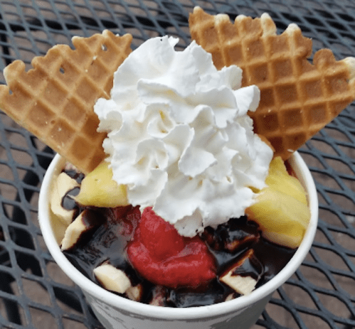 A dessert cup with chocolate sauce, bananas, pineapple, a cherry, whipped cream, and waffle cone pieces on top.