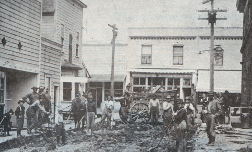 Historic street scene with workers, horses, and a cart in a muddy area, surrounded by buildings and utility poles.