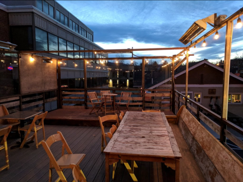 Rooftop patio with wooden tables and chairs, string lights, and a sunset sky in the background.