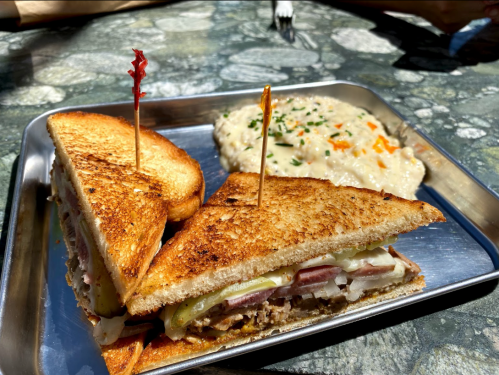 A toasted sandwich with layers of meat and pickles, served on a metal tray alongside a creamy side dish.